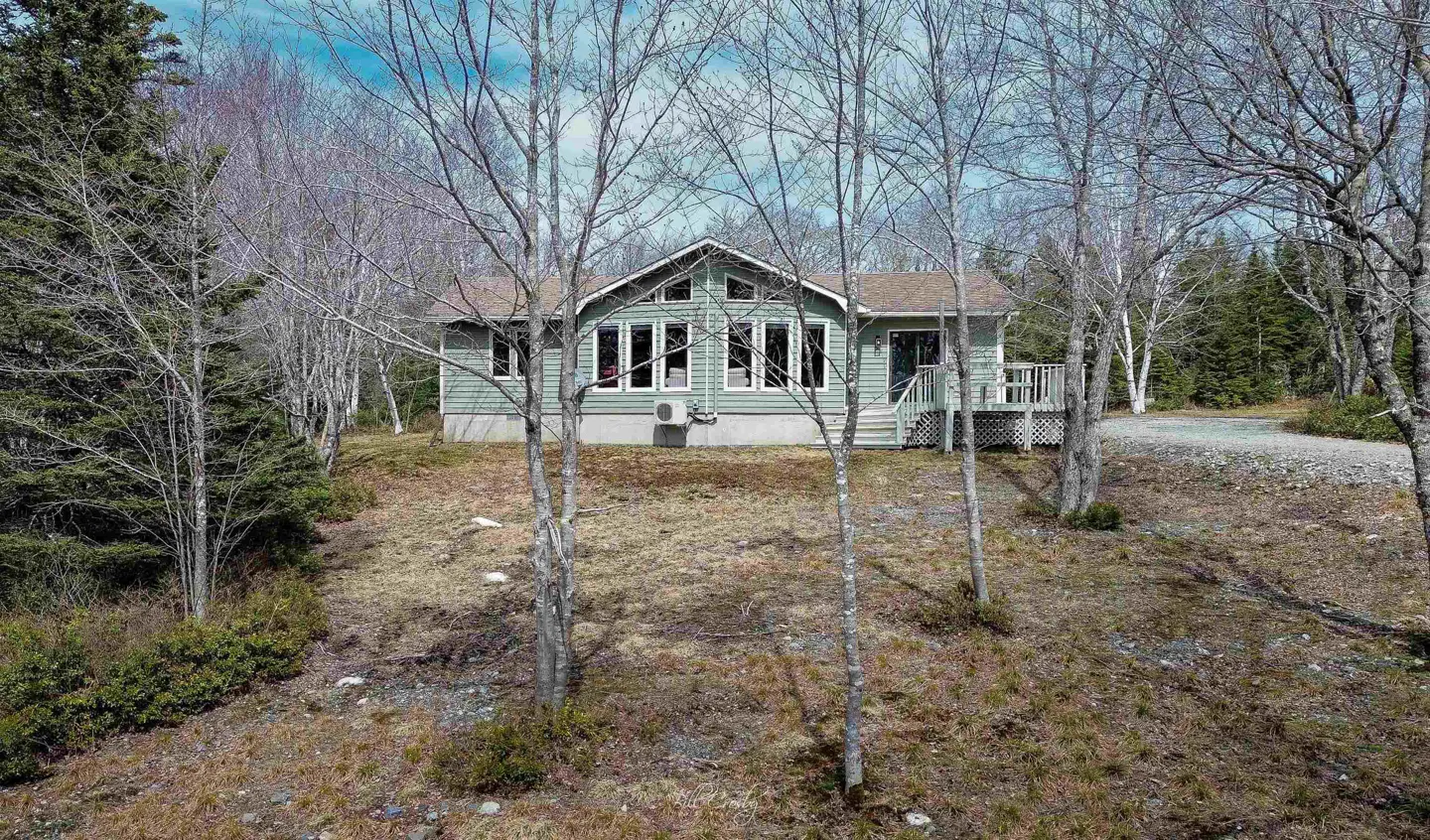 A light green house with a brown roof, surrounded by trees and a grassy lawn. A deck and stairs lead to the front door.