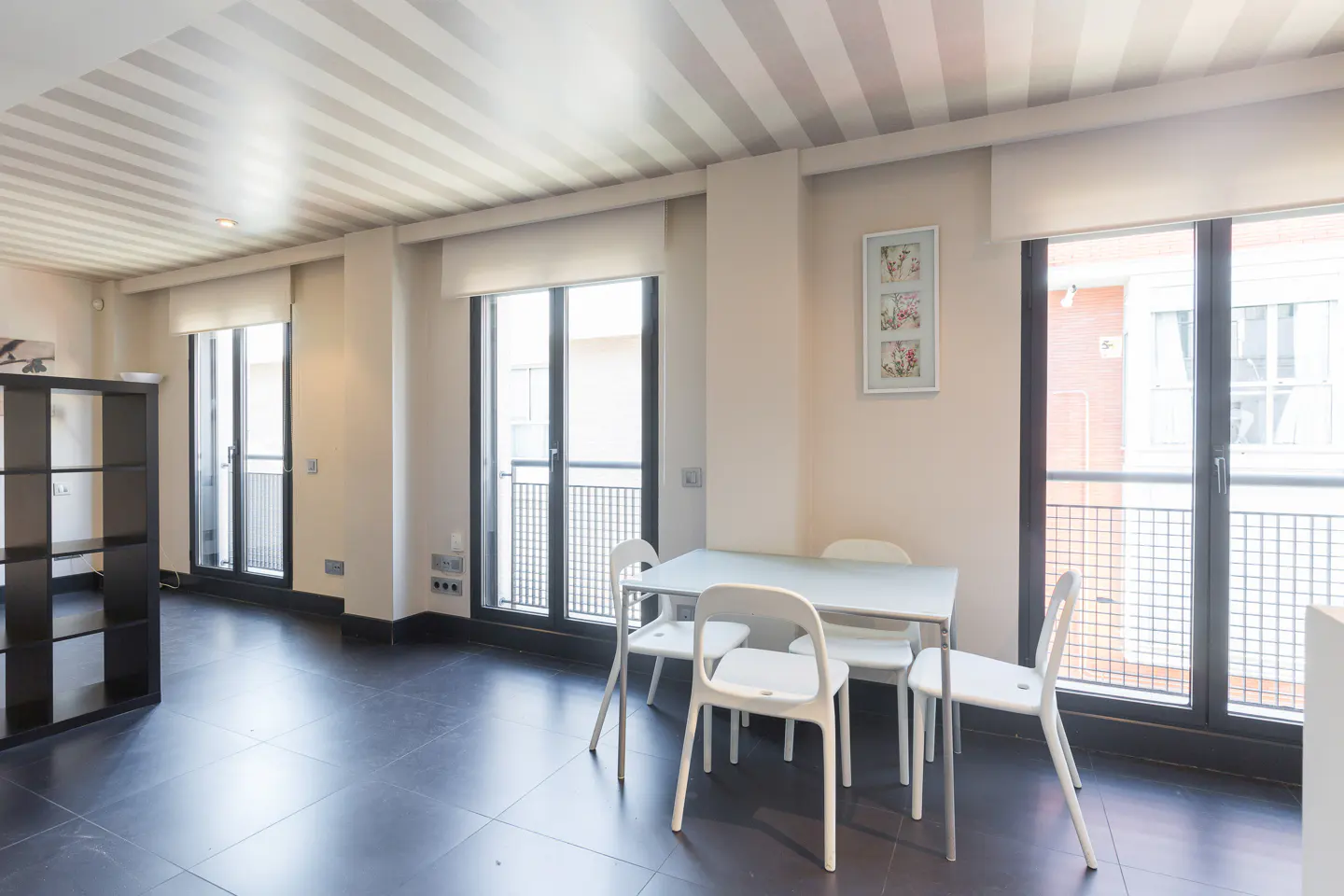 A bright dining area with a white table and chairs, black tile floor, and large windows with balcony views. The ceiling has a striped pattern.