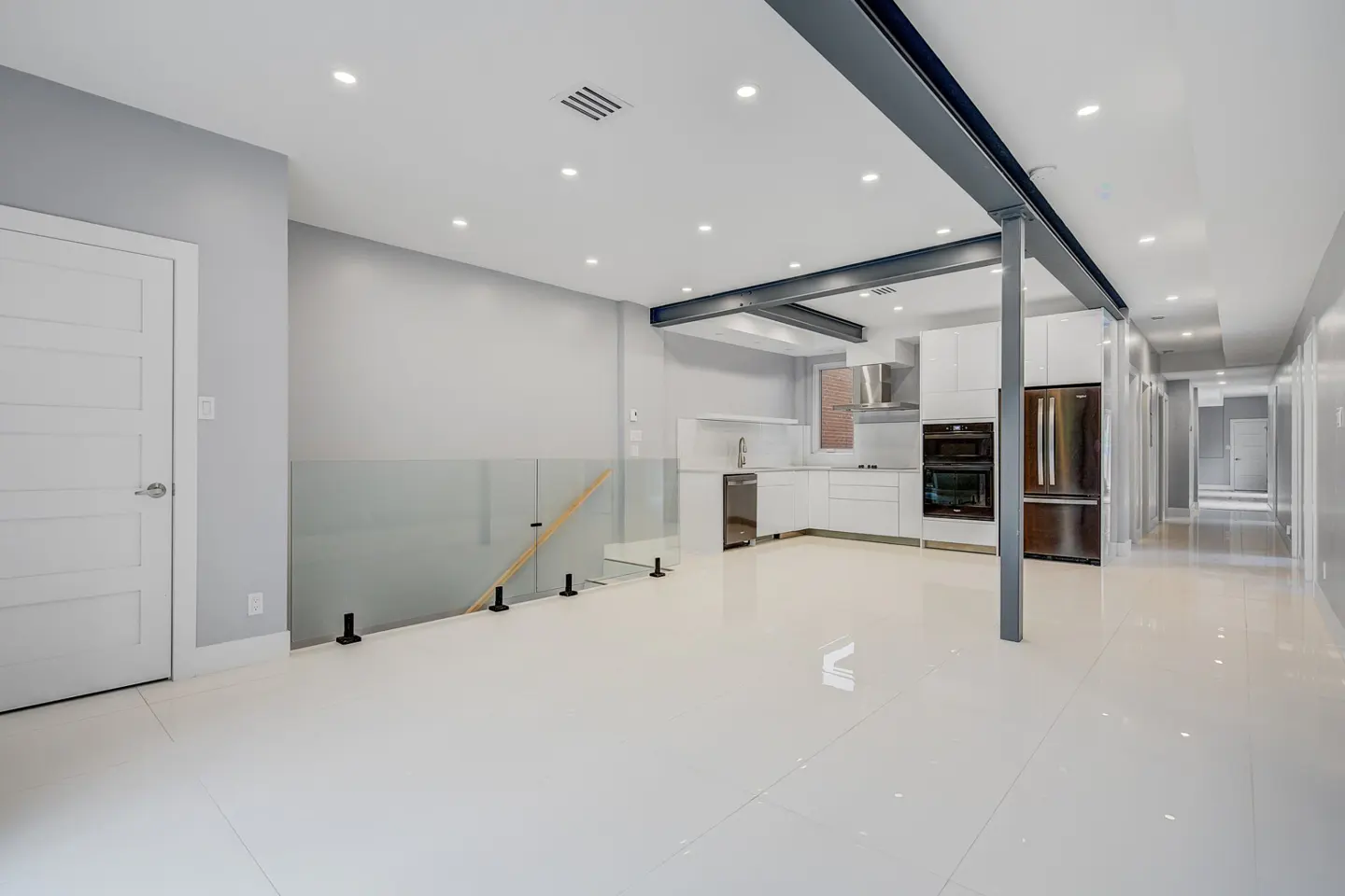 Bright, modern interior with white tile floors, gray walls, and a white kitchen. A glass railing and black steel beams add architectural detail.