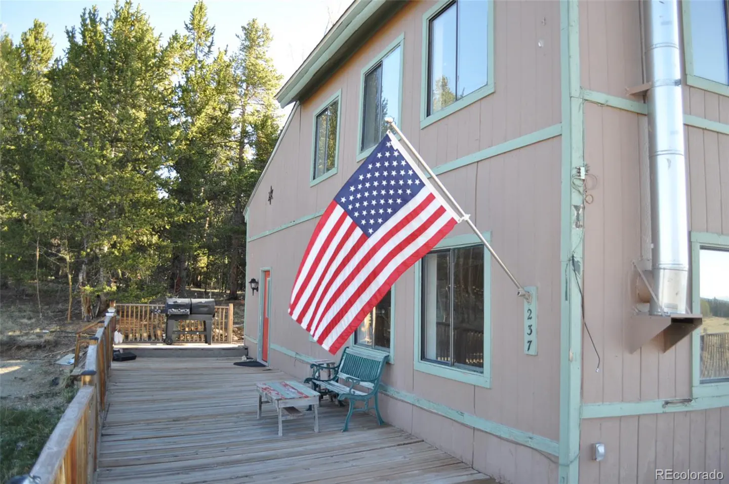 Tan house with green trim, a wooden deck, a green bench, a grill, and an American flag mounted on the exterior wall.