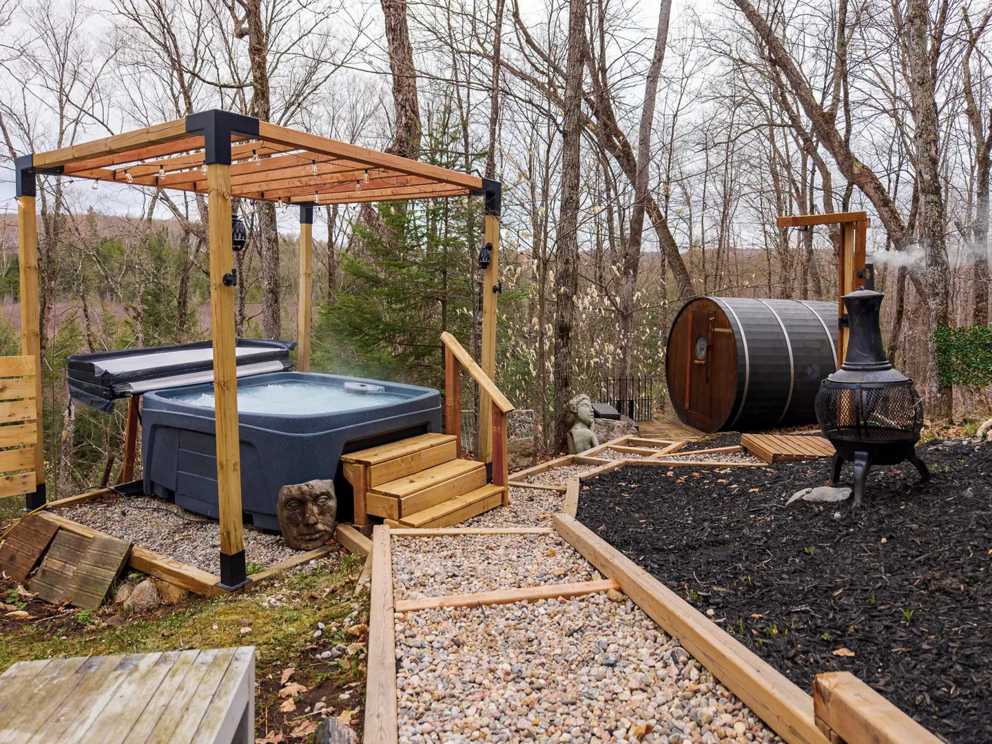 Outdoor spa area with a hot tub under a pergola, a barrel sauna, and a fire pit, surrounded by trees.