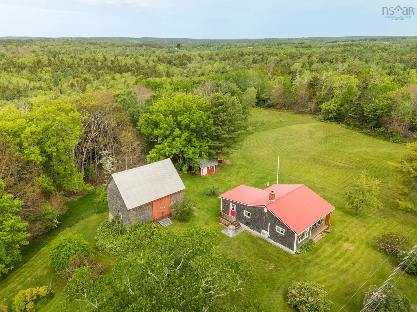 Aerial view of a house with a red roof and a separate barn on a large, grassy lot surrounded by dense forest.