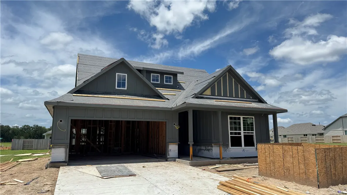 A new gray house under construction with an open garage, unfinished roof, and a concrete driveway under a blue sky.