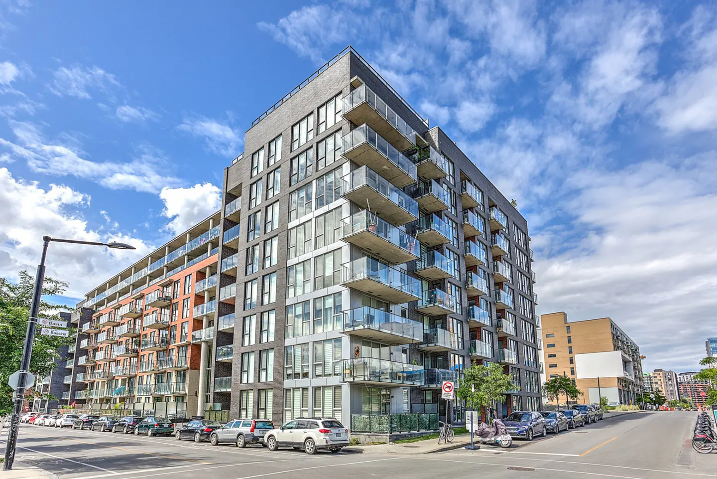 Modern apartment building with glass balconies, parked cars, and a blue sky with clouds.