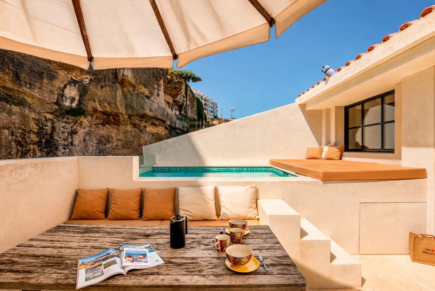 Outdoor patio with a wooden table, cushions, coffee set, and magazine. A small pool and lounge area are visible, with a cliffside backdrop.