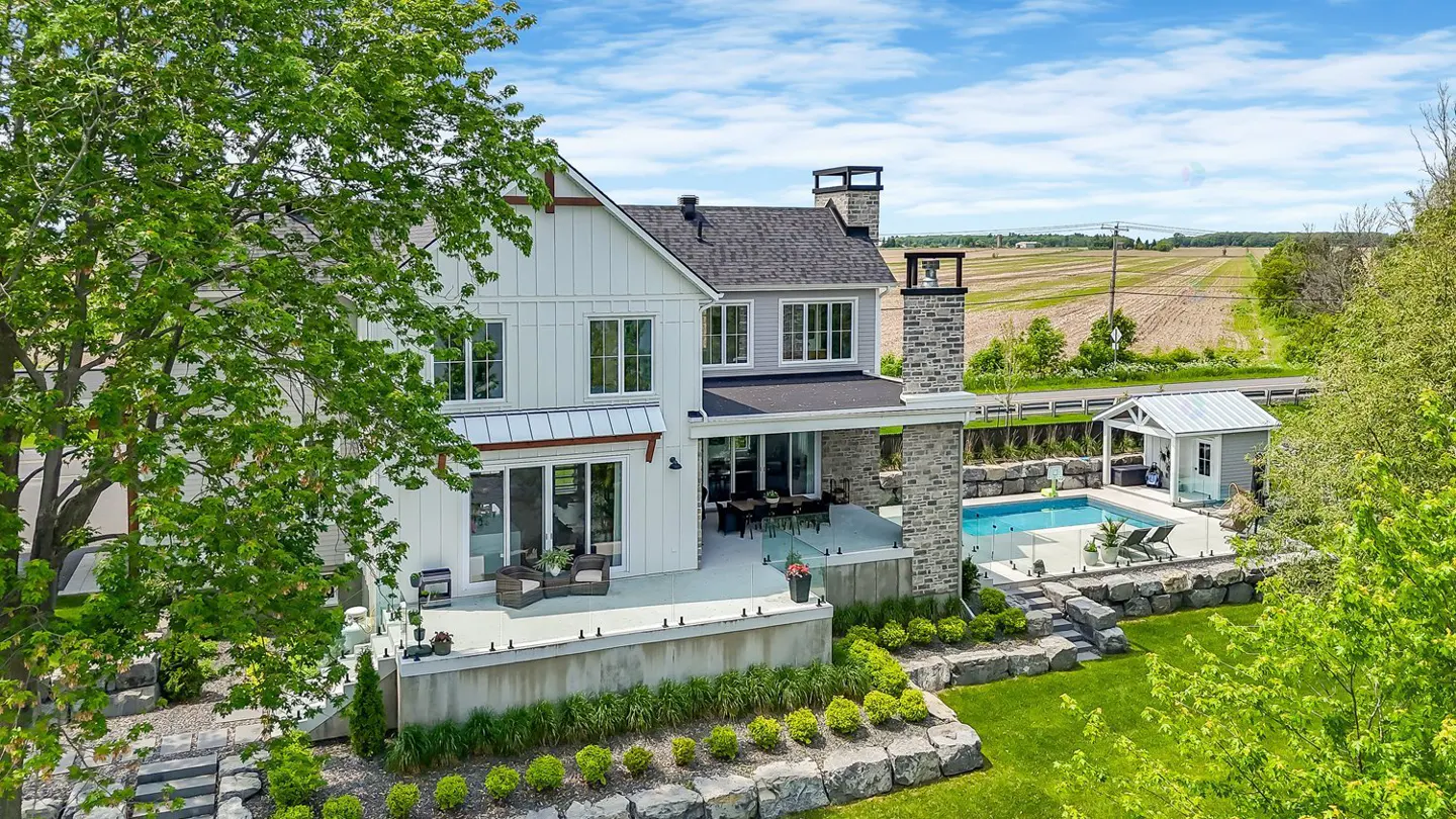 Aerial view of a modern farmhouse with a pool, patio, and stone landscaping. A field is visible in the background.