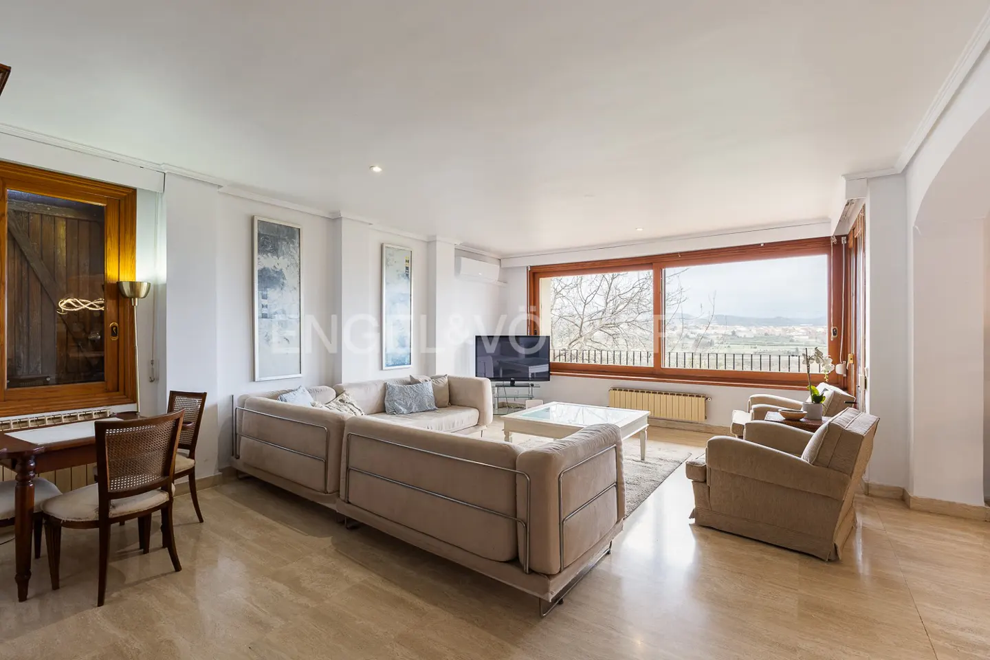 Bright living room with beige sectional sofa, two armchairs, and a large window with a view. A dining table sits to the left.