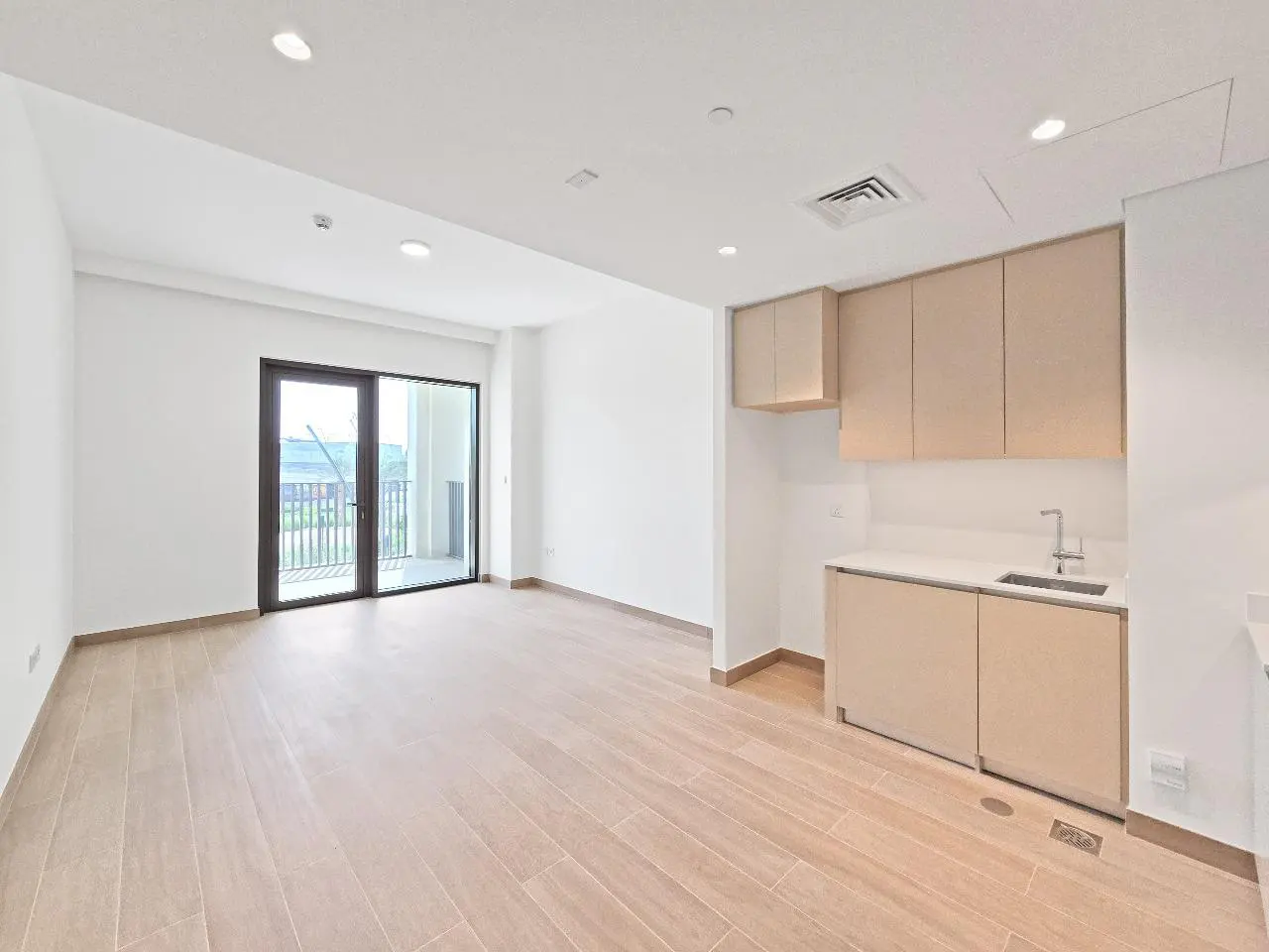Bright, empty apartment with light wood floors, white walls, and a small kitchen area with tan cabinets. A glass door leads to a balcony.