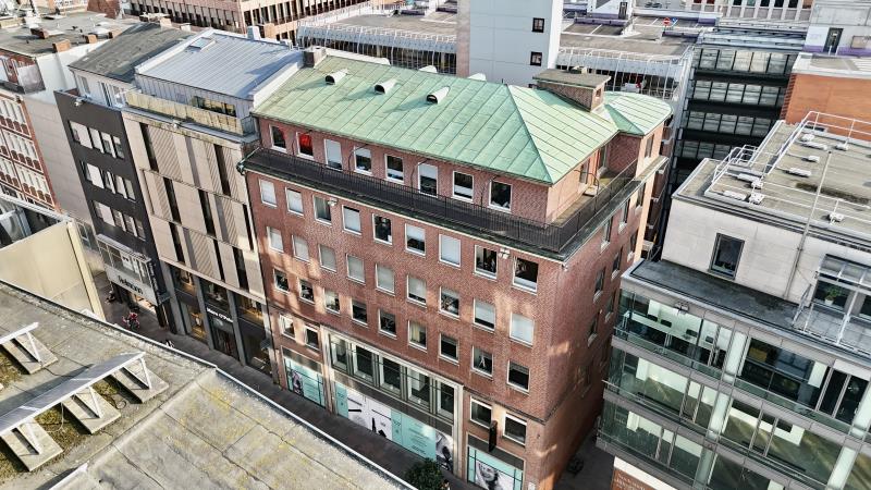 High-angle view of a red brick building with a green metal roof surrounded by modern urban architecture.