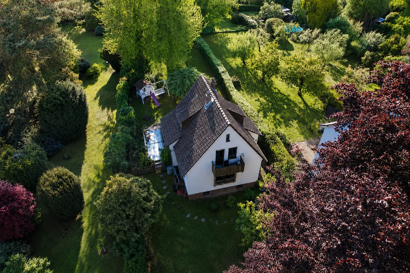 Aerial view of a white house with a brown roof, surrounded by green trees and lawn. A children's playhouse is visible in the background.