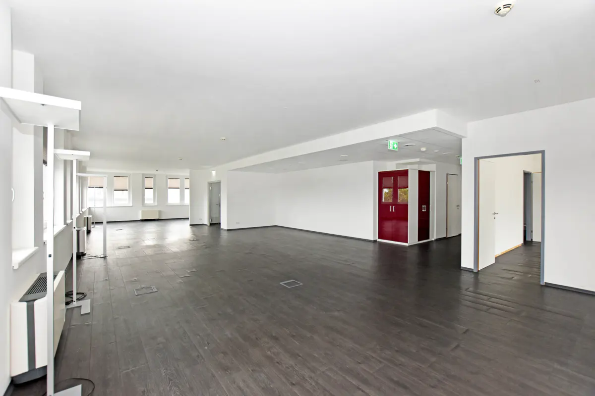 Bright, empty office space with dark wood floors, white walls, and windows. A red cabinet stands near a doorway.