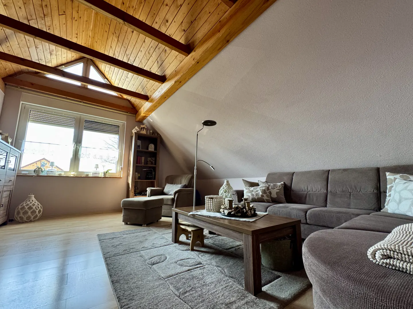 Attic living room with wood-beamed ceiling, brown sectional sofa, coffee table, and rug. Window with blinds lets in natural light.