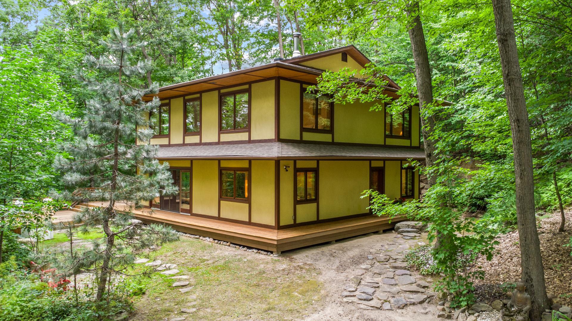 Two-story yellow house with brown trim surrounded by lush green trees and a stone path leading to the entrance.