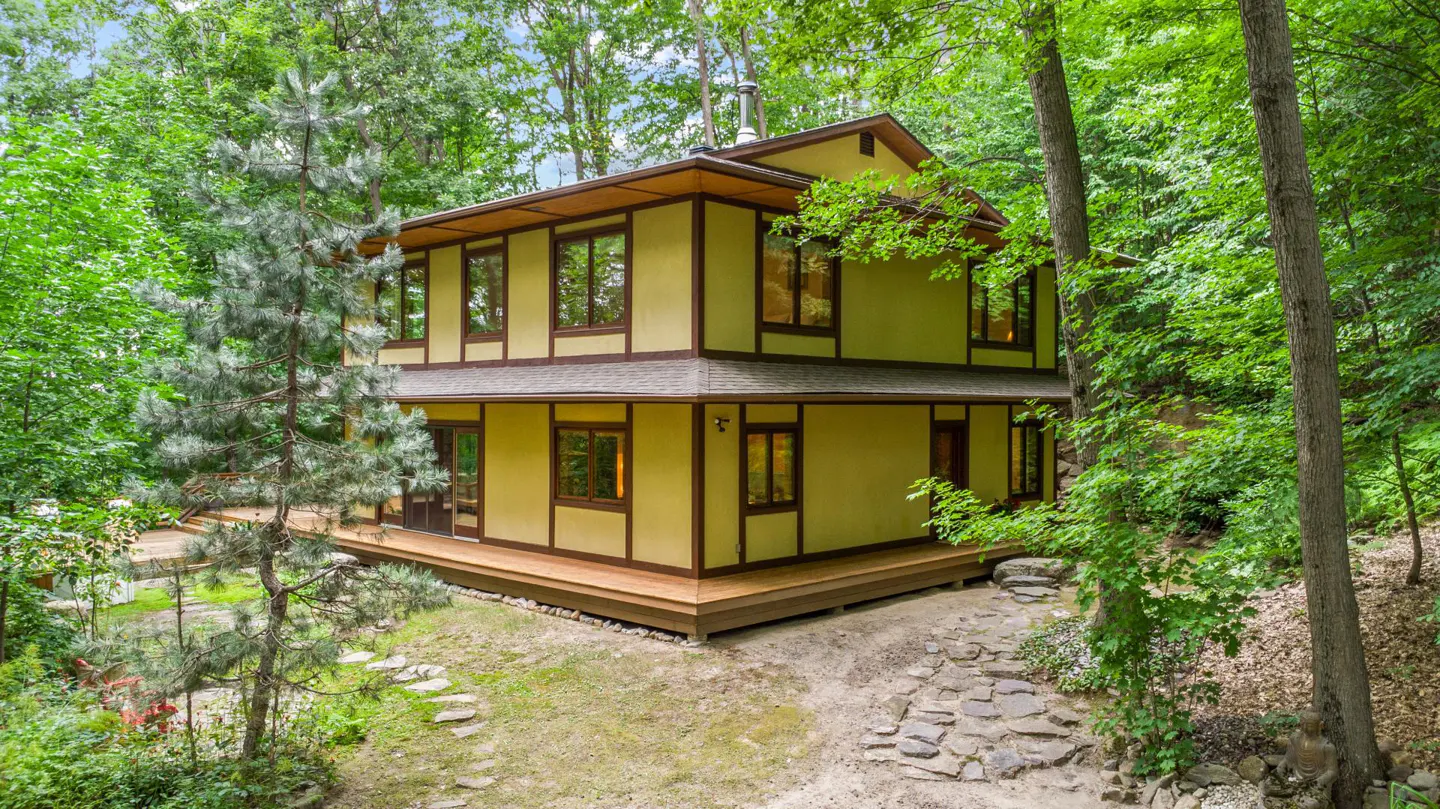 Two-story yellow house with brown trim surrounded by lush green trees and a stone path leading to the entrance.