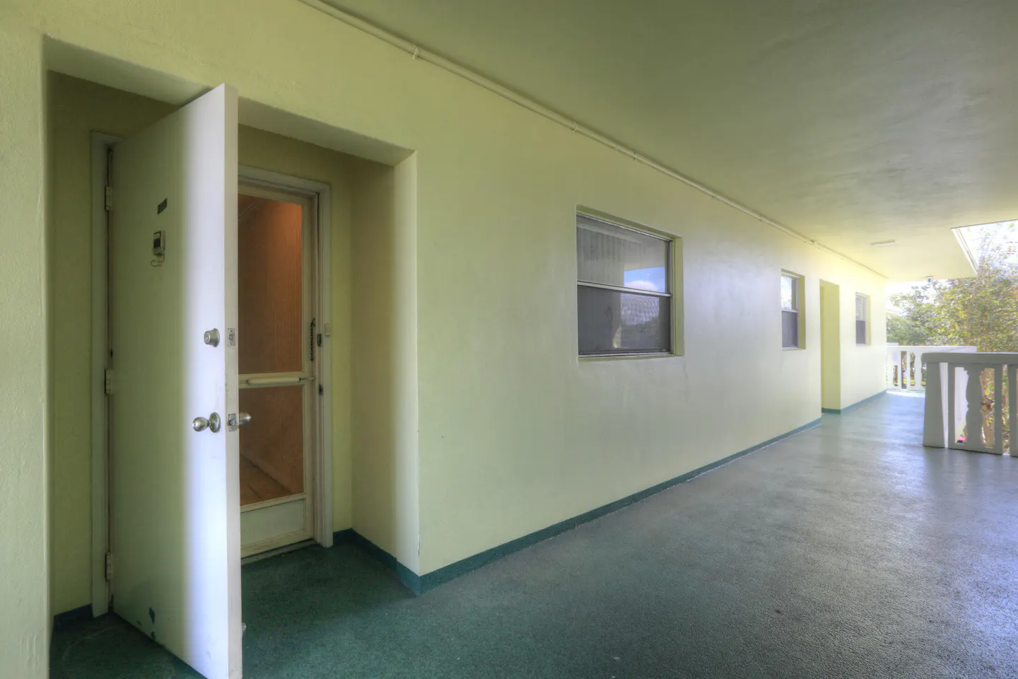 Exterior view of a light yellow apartment building hallway with an open white door. Windows line the hallway, and a white railing is visible at the end.