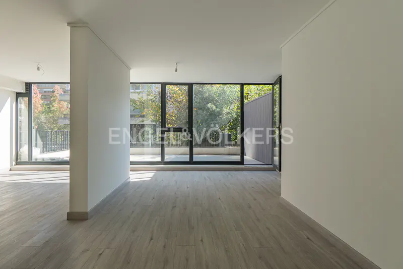 Bright, empty room with light wood floors and white walls. Large windows offer views of trees and a balcony.
