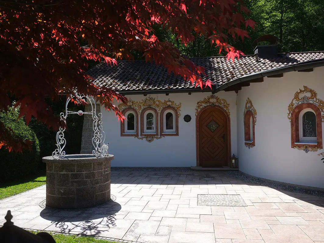 Exterior view of a white building with a tiled roof, decorative windows, and a wooden door. A stone well sits in the foreground, framed by red leaves.