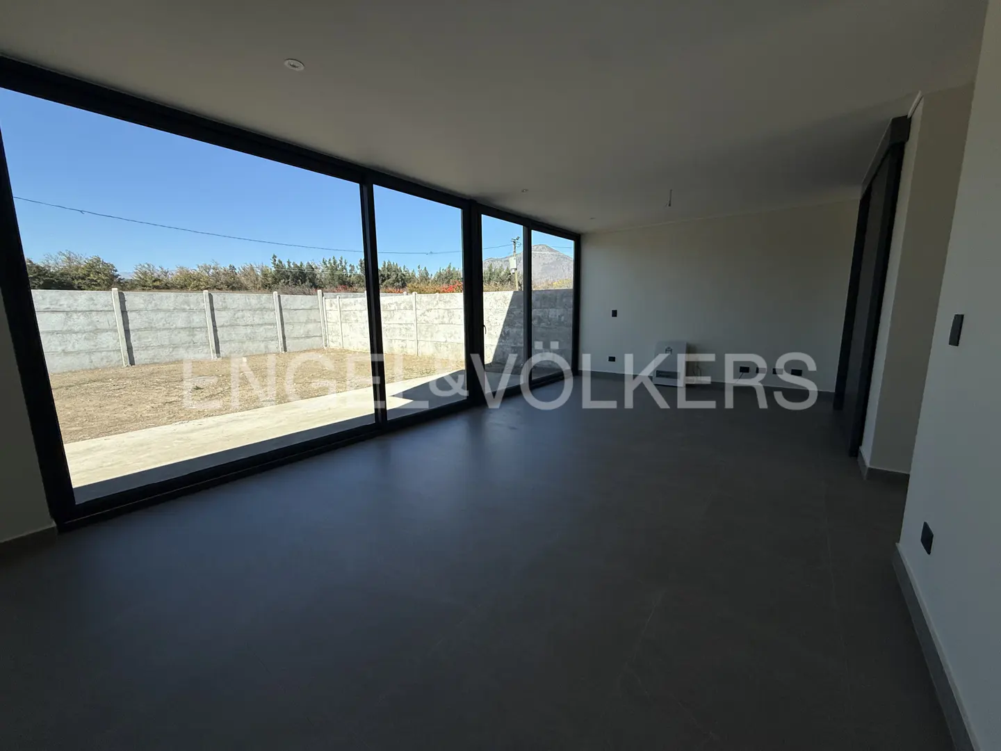 An empty room with gray floors and white walls has large windows looking out to a yard with a concrete fence.