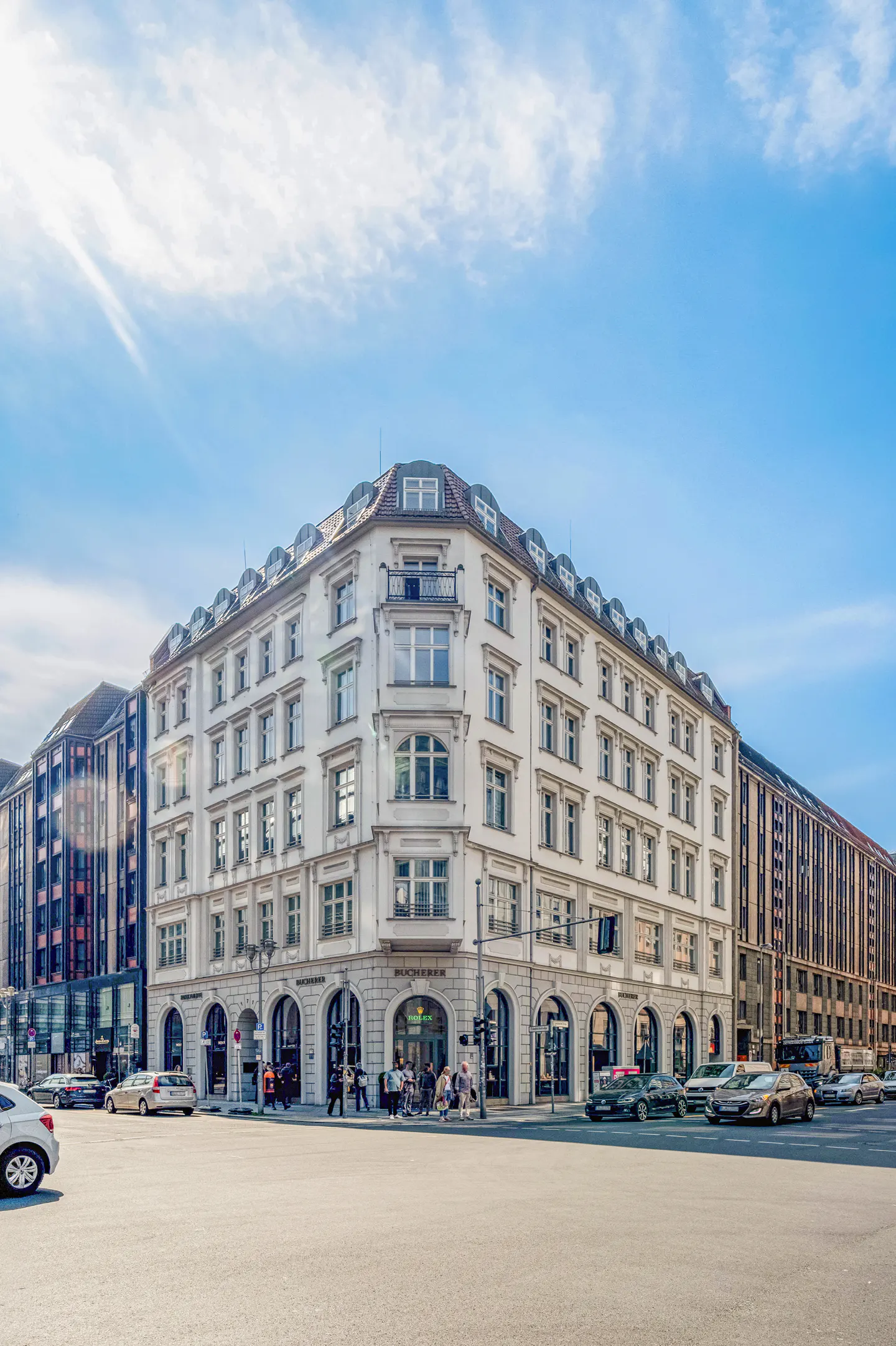 Street view of a white, multi-story building with shops on the ground floor, cars, and pedestrians on a sunny day.