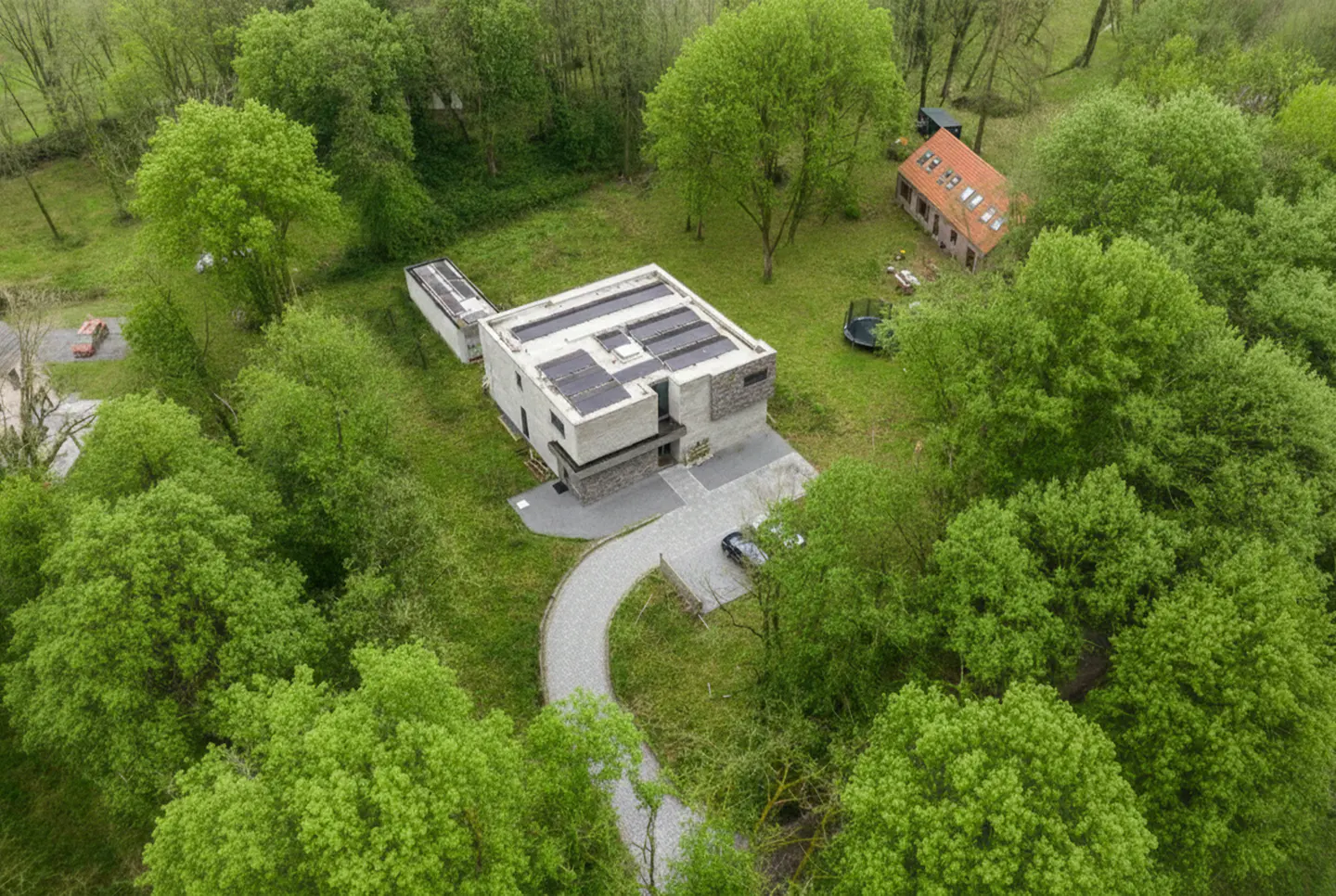 Aerial view of a modern, light gray house with solar panels, surrounded by lush green trees and a winding driveway.