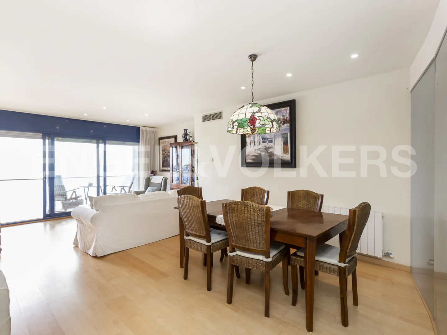 Bright, open-plan living and dining area with wood floors. A dark wood dining table with six chairs sits under a stained-glass pendant light. A white sofa faces large windows.