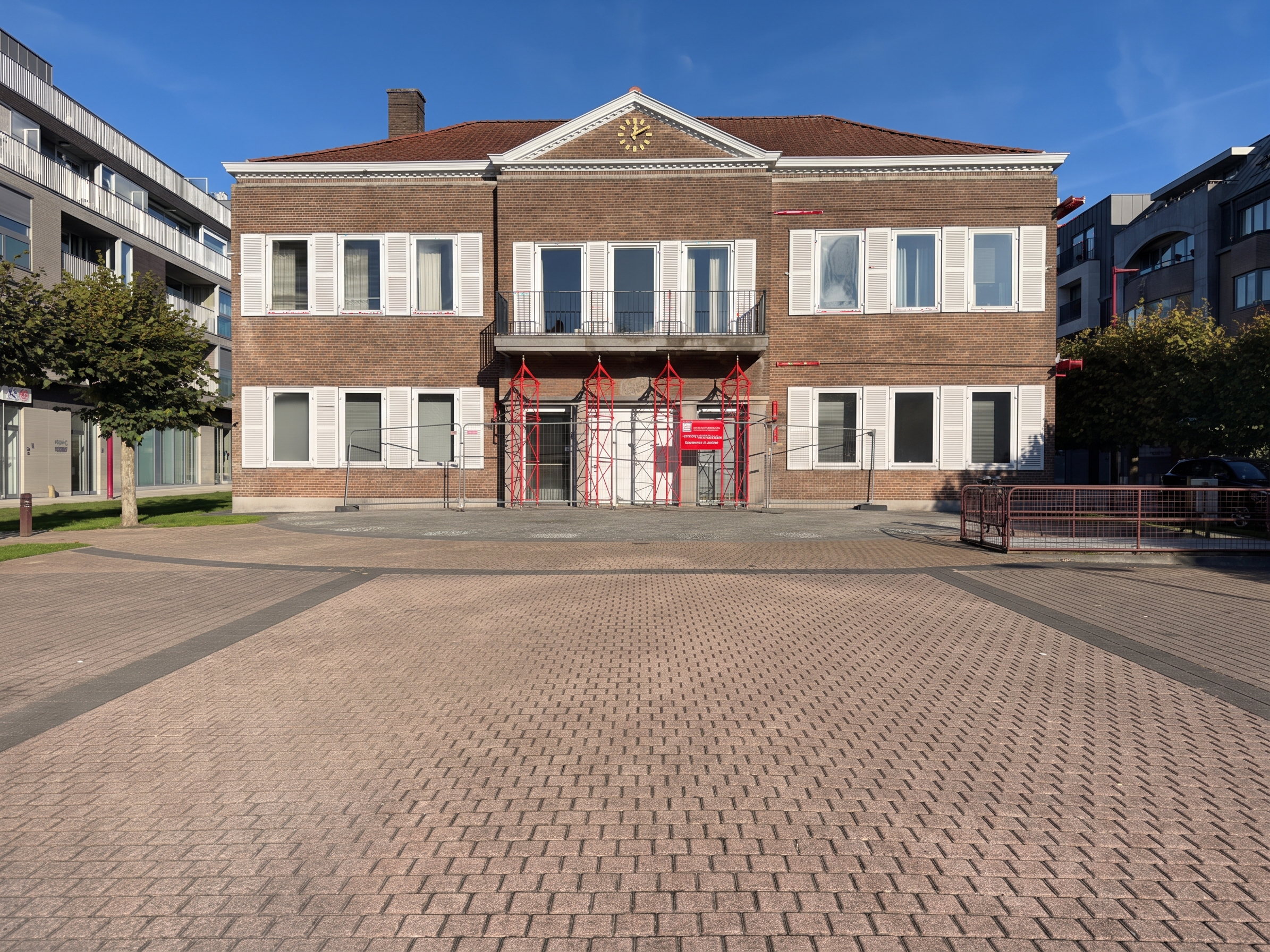 A historic brown brick building with white shutters and a central balcony, featuring red scaffolding in front of the entrance. [AI-generated text]