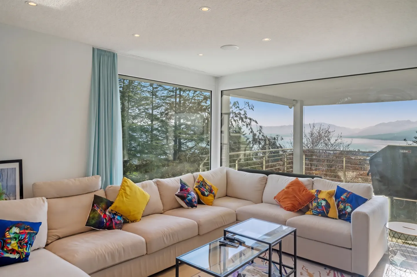 Bright living room with a beige sectional sofa, colorful pillows, and glass tables. Large windows offer views of trees, a lake, and mountains.