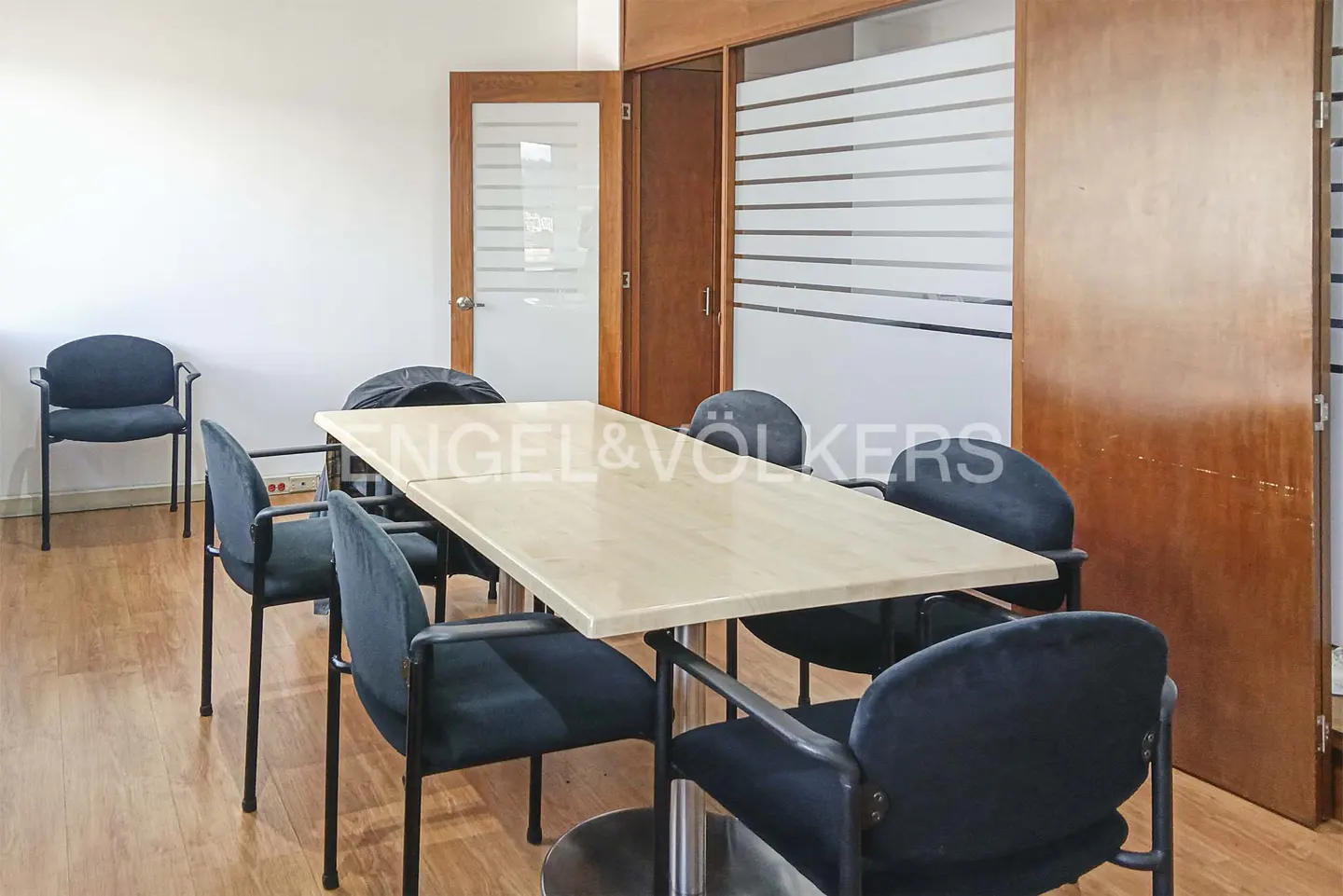 Conference room with a light wood table, black chairs, and wood floors. A glass door and frosted glass wall are in the background.