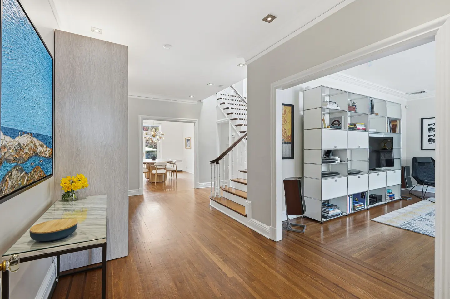 Bright foyer with hardwood floors, white walls, and a spiral staircase. A blue ocean painting hangs above a marble table with yellow flowers.