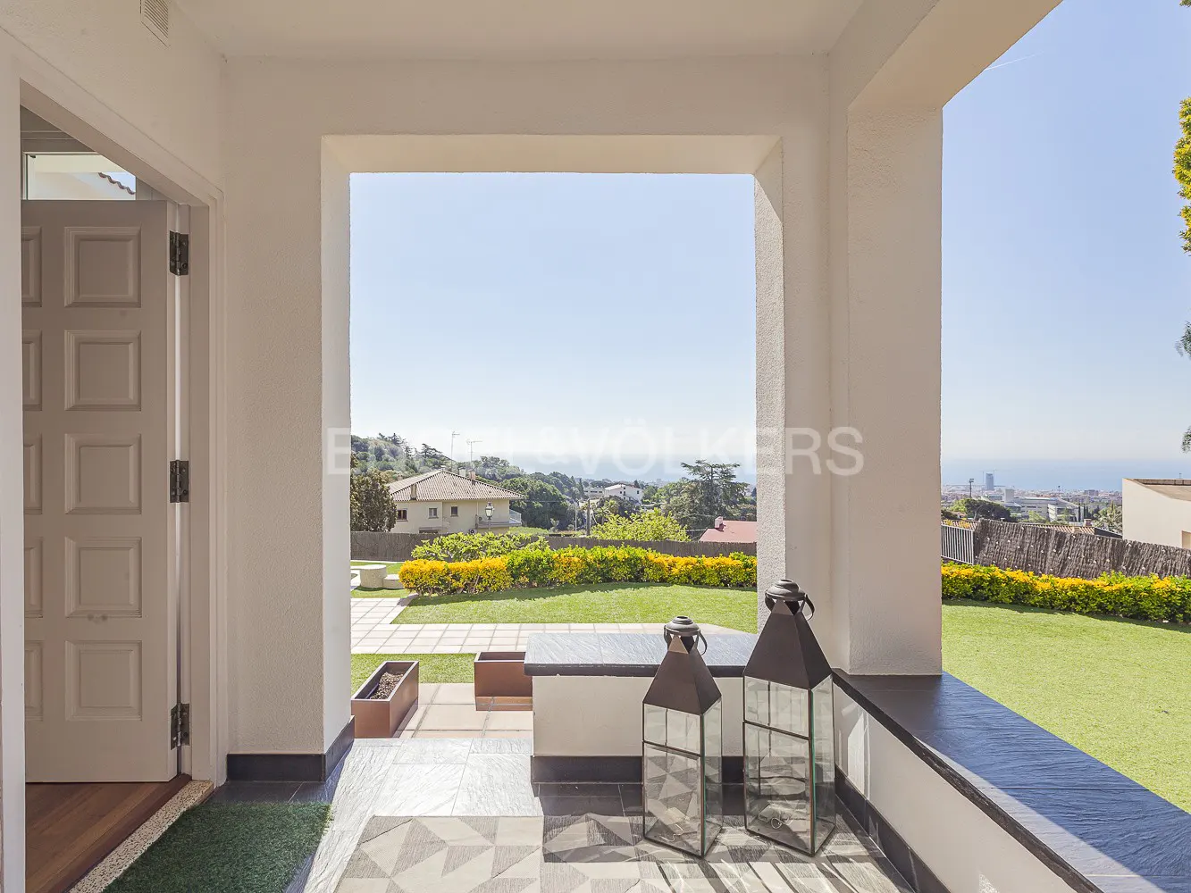 View from a white-columned porch with lanterns, overlooking a green lawn, houses, and the sea under a blue sky.