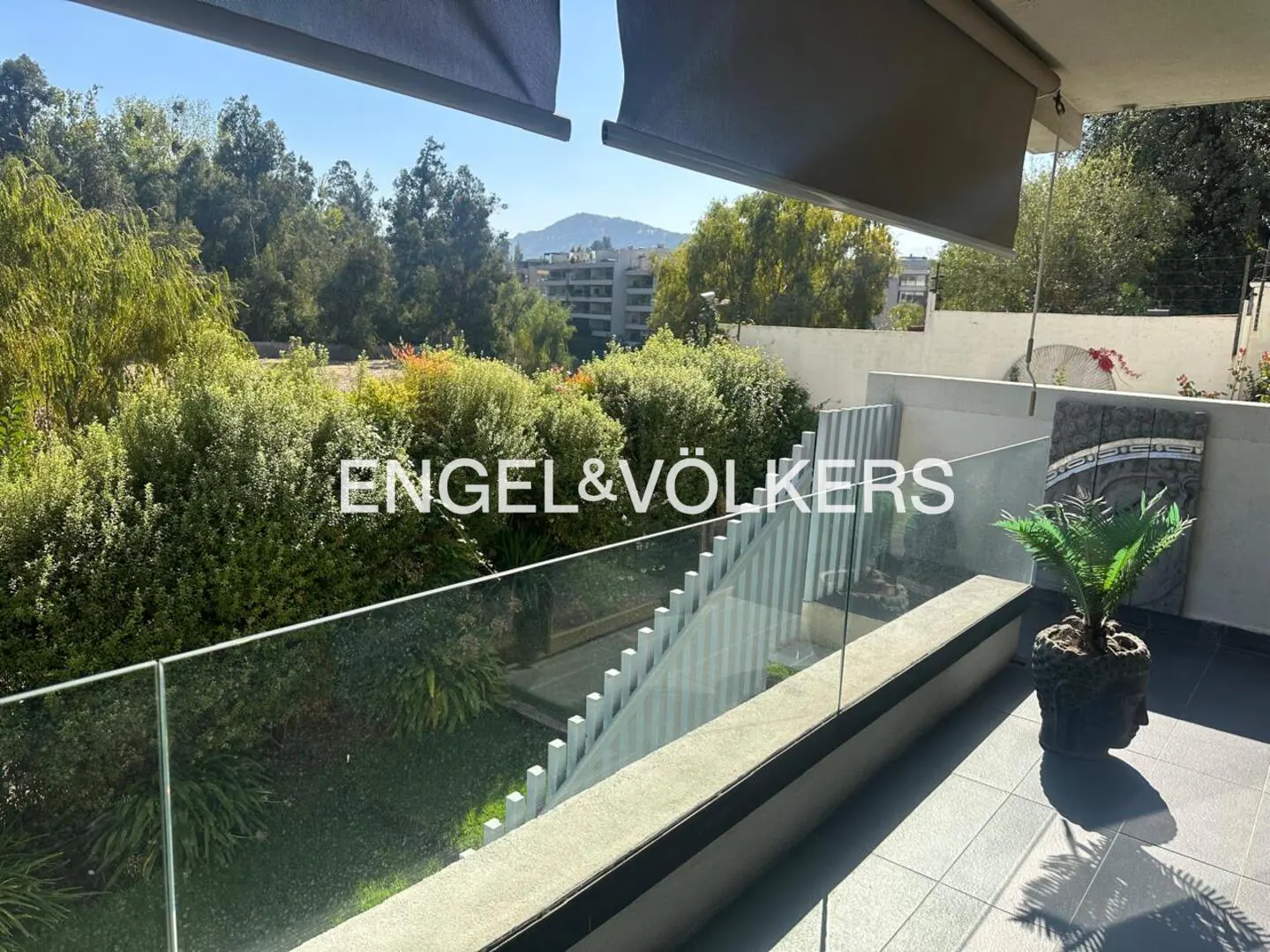 Balcony view with glass railing, overlooking lush greenery and distant buildings under a blue sky. A potted plant sits on the tiled floor.