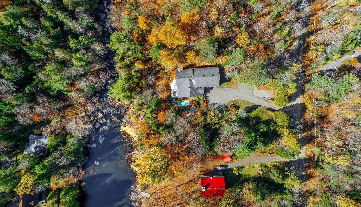 Aerial view of a house with a gray roof surrounded by colorful autumn trees and a river.