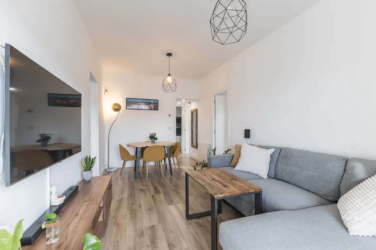 Bright living room with gray sectional sofa, wood coffee table, dining table with yellow chairs, and a large TV on the wall.