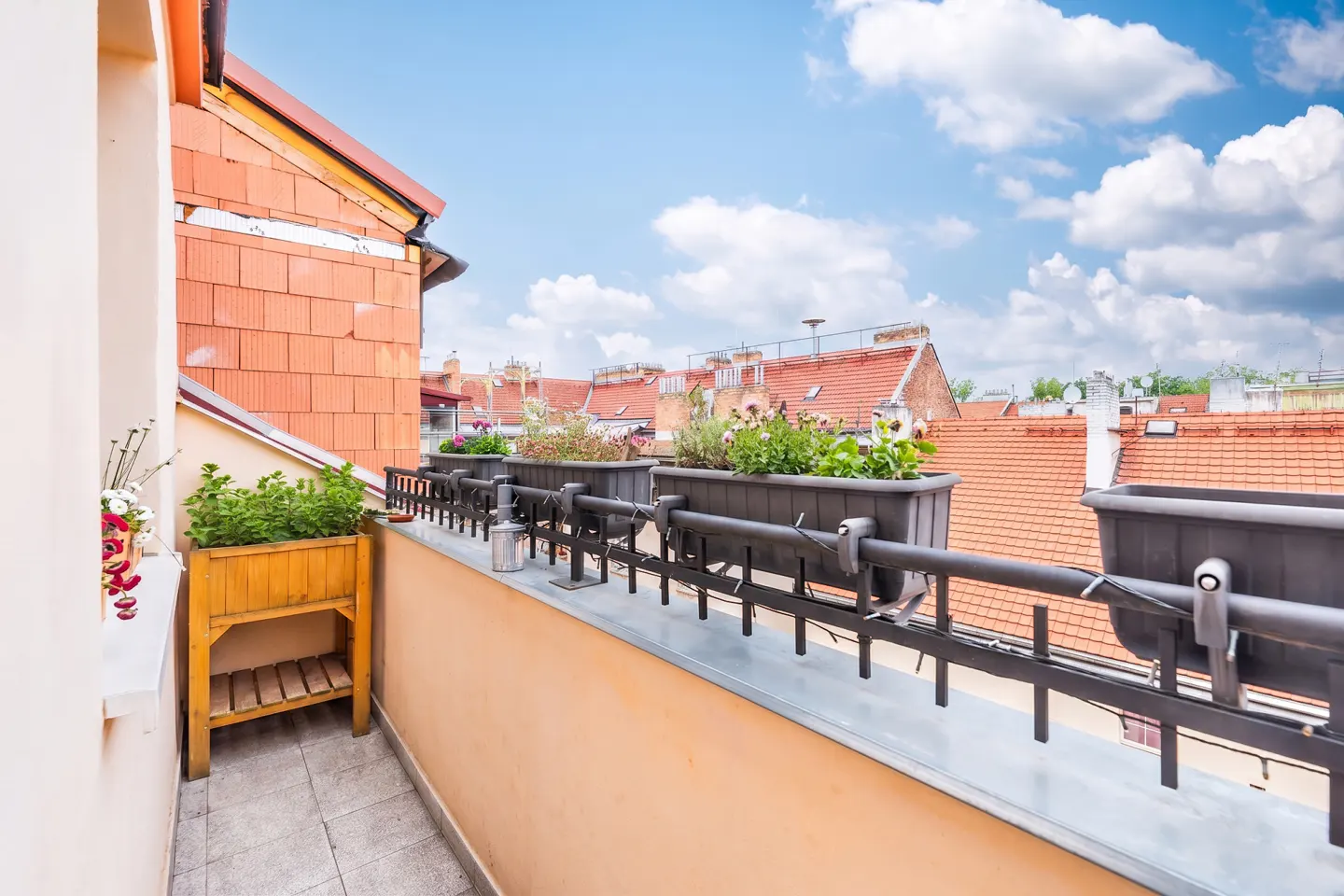 A balcony with a wooden planter and railing flower boxes overlooking a city skyline with orange tiled roofs under a blue sky.