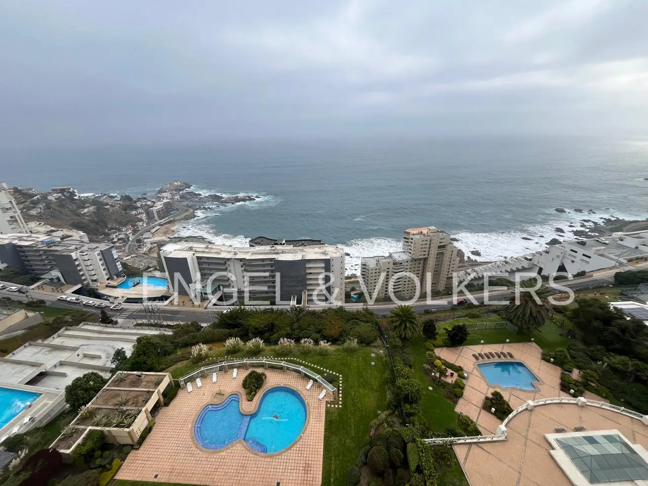 High-angle view of coastal apartment buildings with swimming pools, lush green landscaping, and a cloudy ocean horizon.