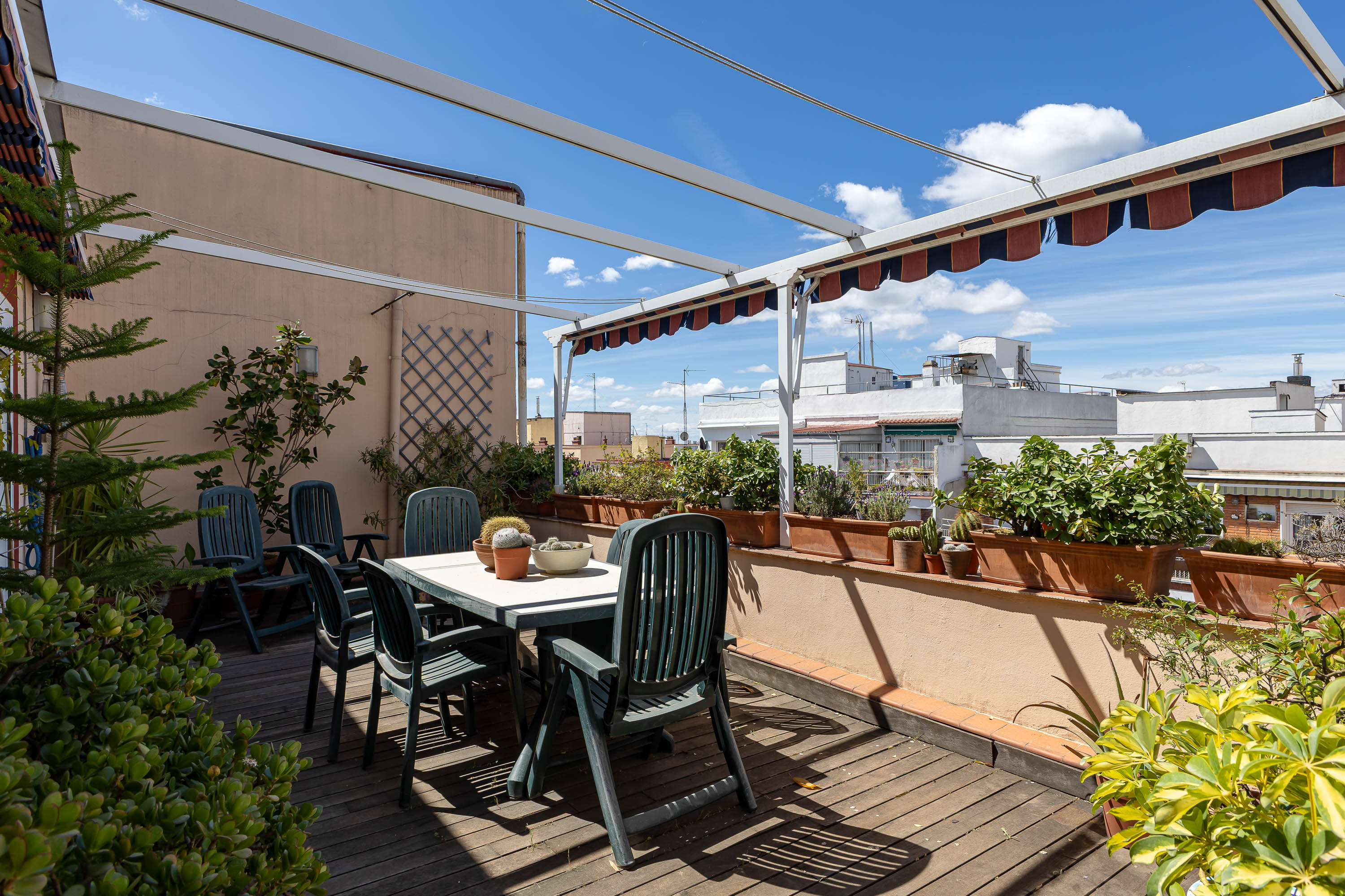 Rooftop patio with a white table, green chairs, and potted plants. Awnings provide shade, and the sky is blue with clouds.