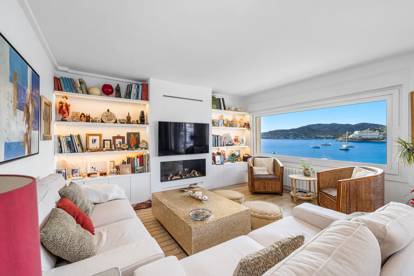 Bright living room with white sofas, woven coffee table, and built-in shelves. Large window shows a blue ocean view with boats and green hills.