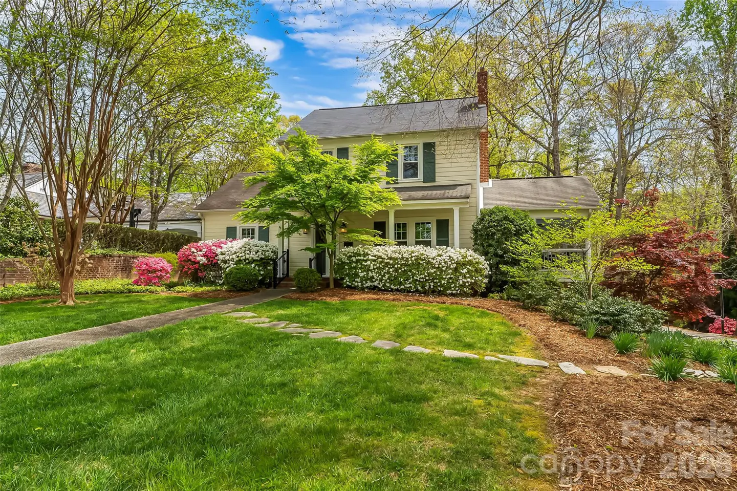 Two-story beige house with green shutters, surrounded by green trees and colorful bushes. A stone walkway leads to the front door.
