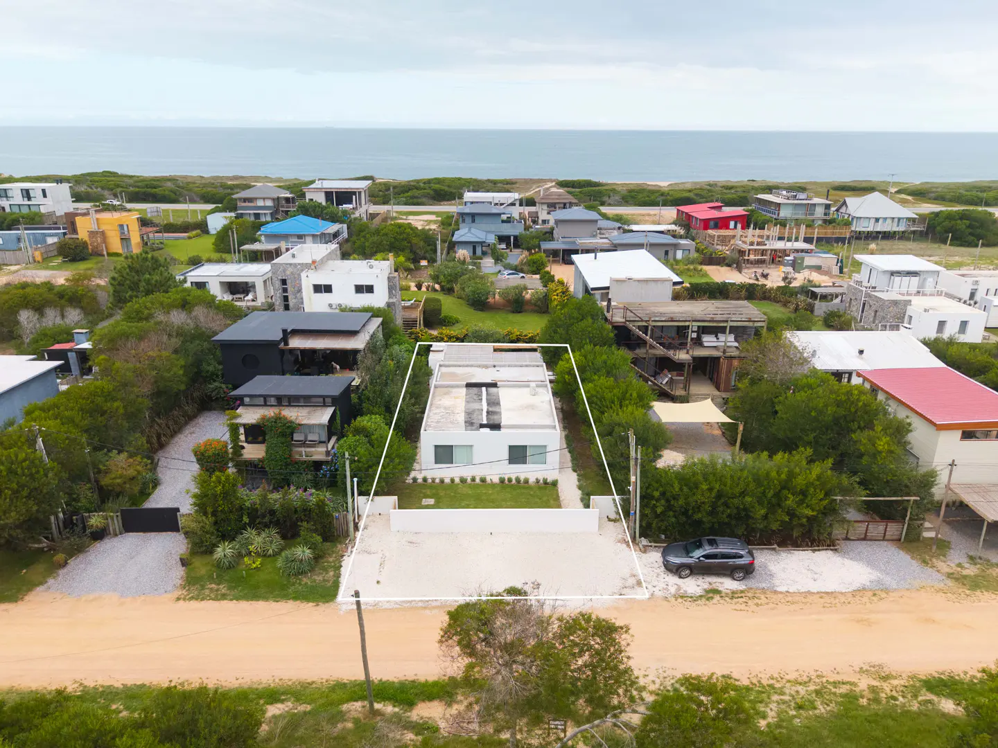Aerial view of a modern white house with a flat roof, surrounded by green trees and other colorful houses near the ocean.