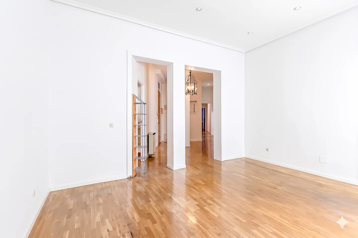 Empty room with white walls, hardwood floors, and two doorways leading to hallways. A chandelier hangs in the right doorway.