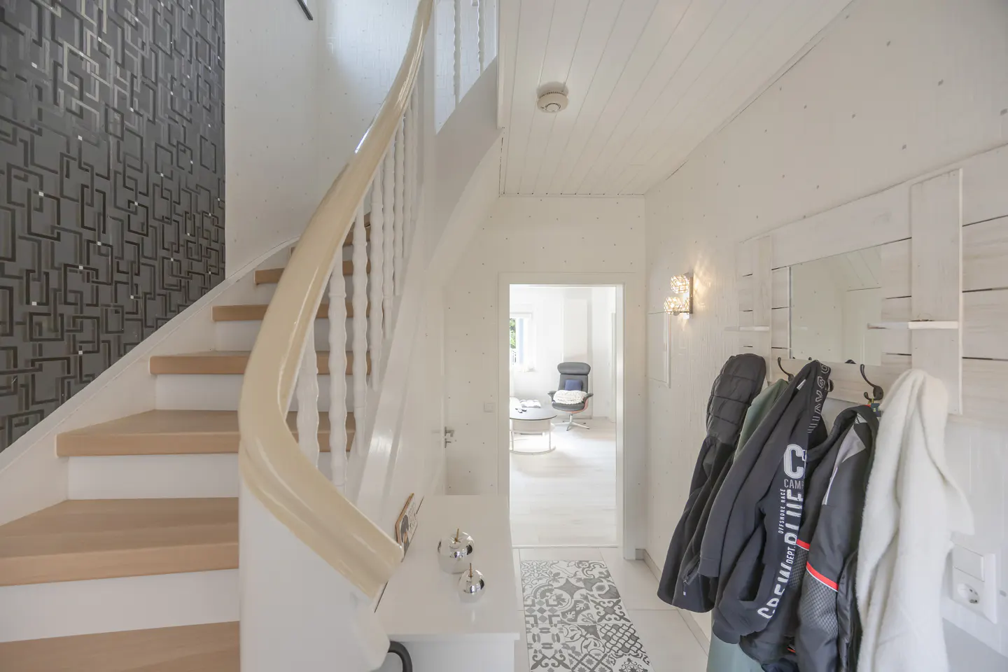 Bright entryway with a staircase, white walls, and a coat rack with jackets. A doorway leads to a living room with a chair.