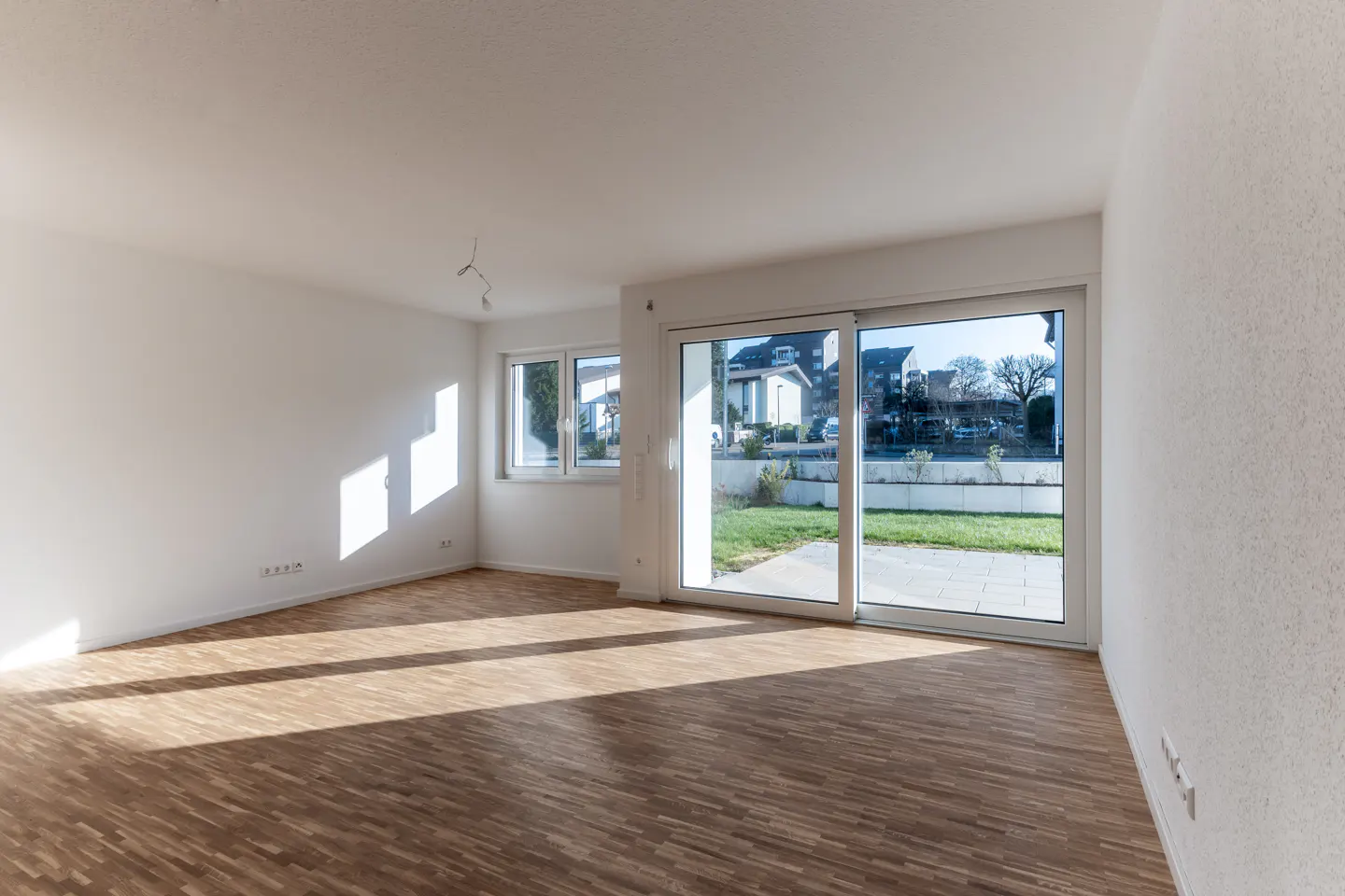 Bright, empty room with wood floors, white walls, and large sliding glass doors to a backyard. Sunlight streams in.