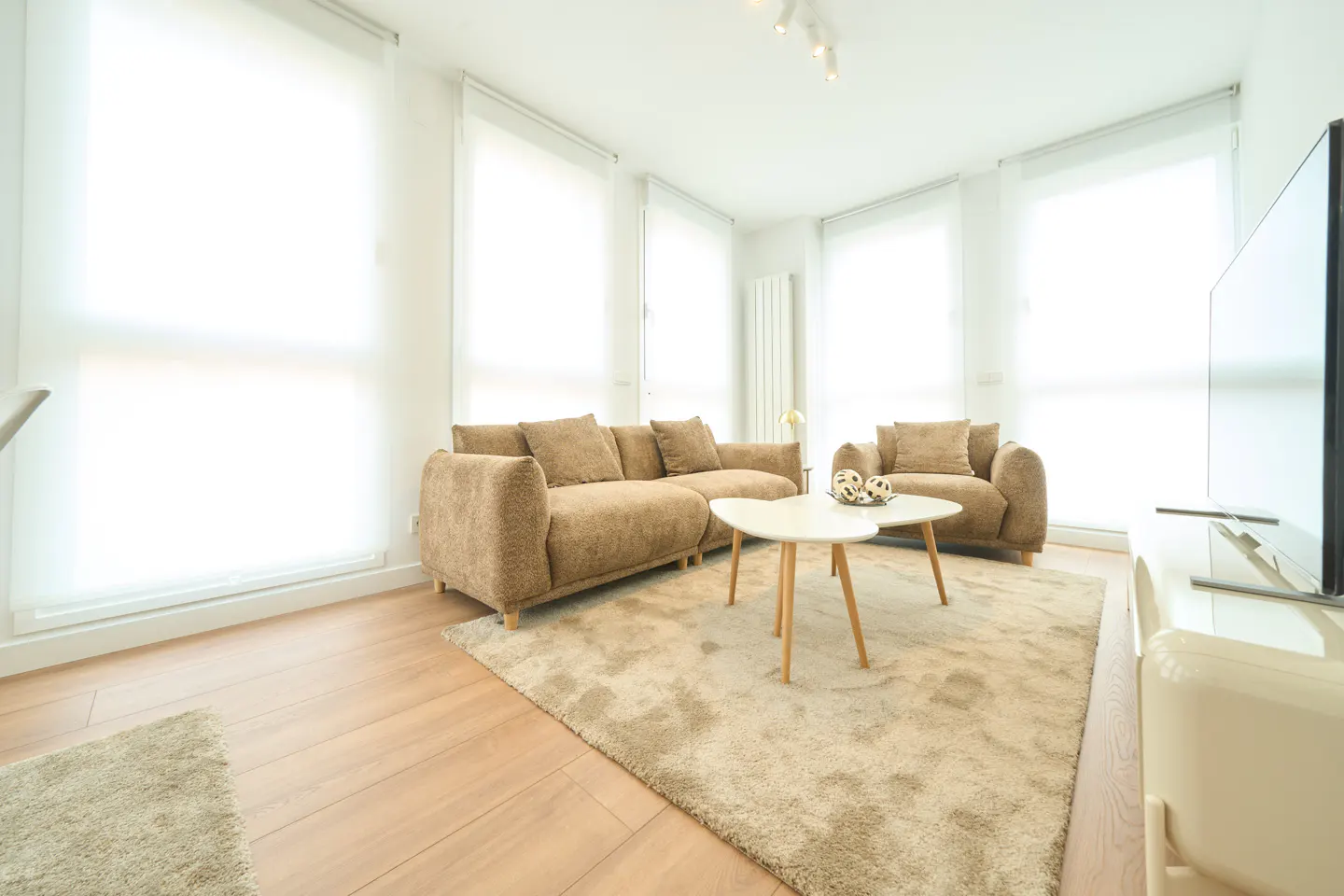 Bright living room with a brown sofa, armchair, and a white coffee table on a beige rug. Large windows with white blinds.