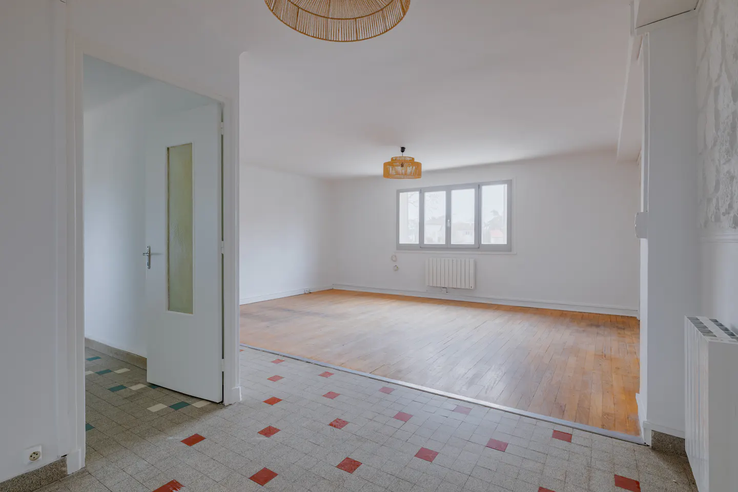 A bright, empty room with hardwood floors and white walls is viewed through an open door. A window and radiator are visible.
