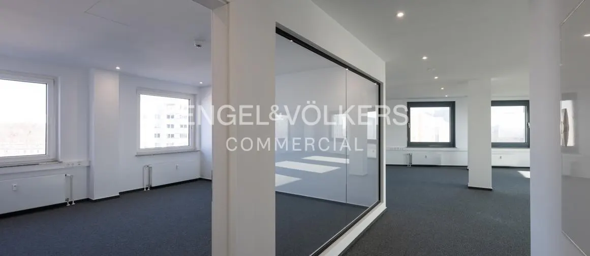 Modern, minimalist office space with white walls, dark gray carpet, large windows, and a glass partition wall.