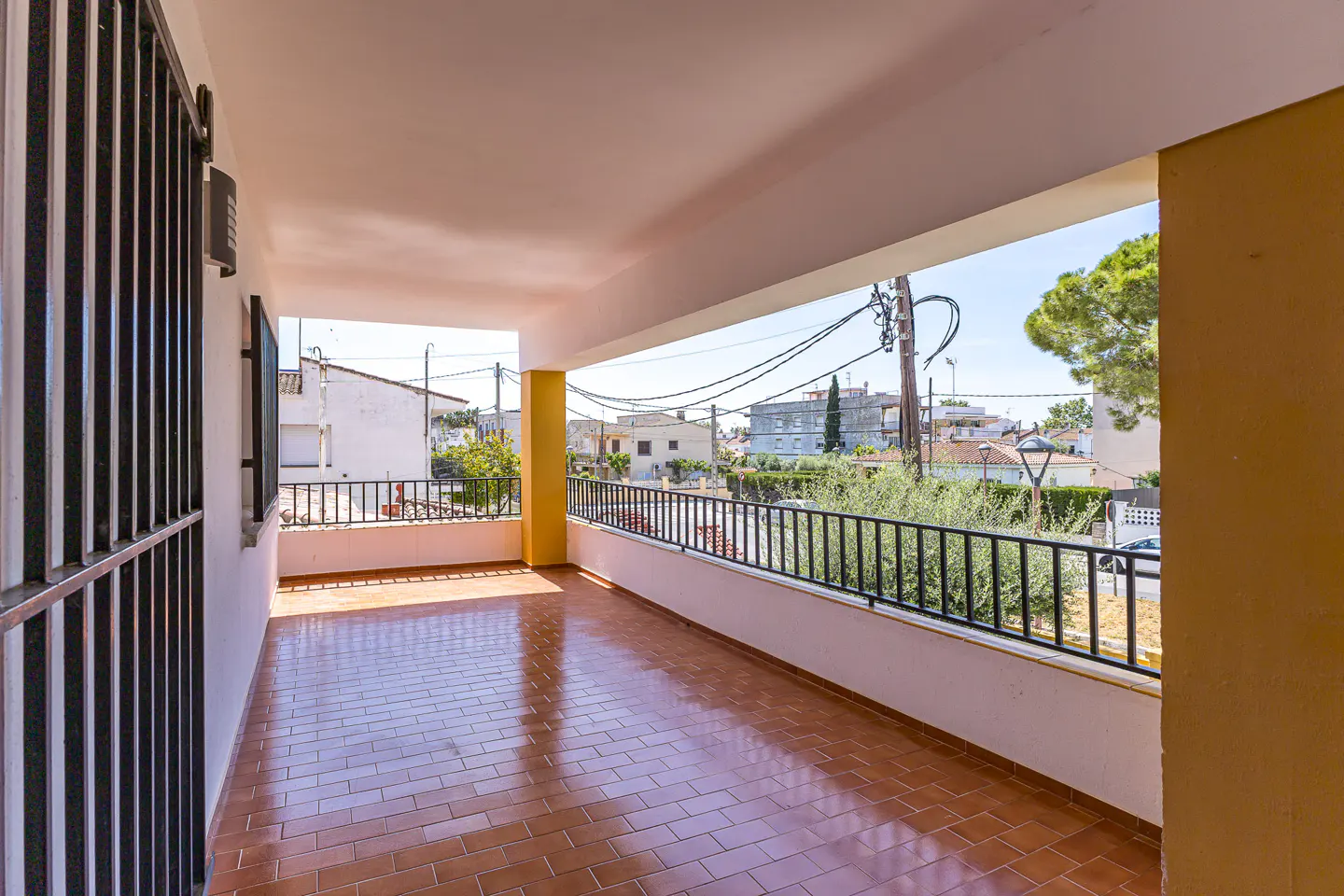 Covered balcony with brown tile floor and black metal railing. View of neighborhood with houses and trees. Yellow support column.