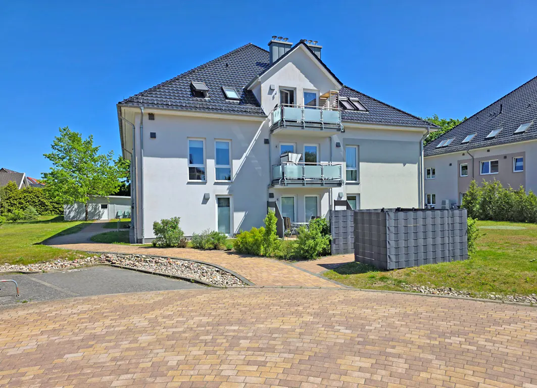 Modern three-story apartment building with white walls, dark roof, and balconies, set in a sunny residential area.