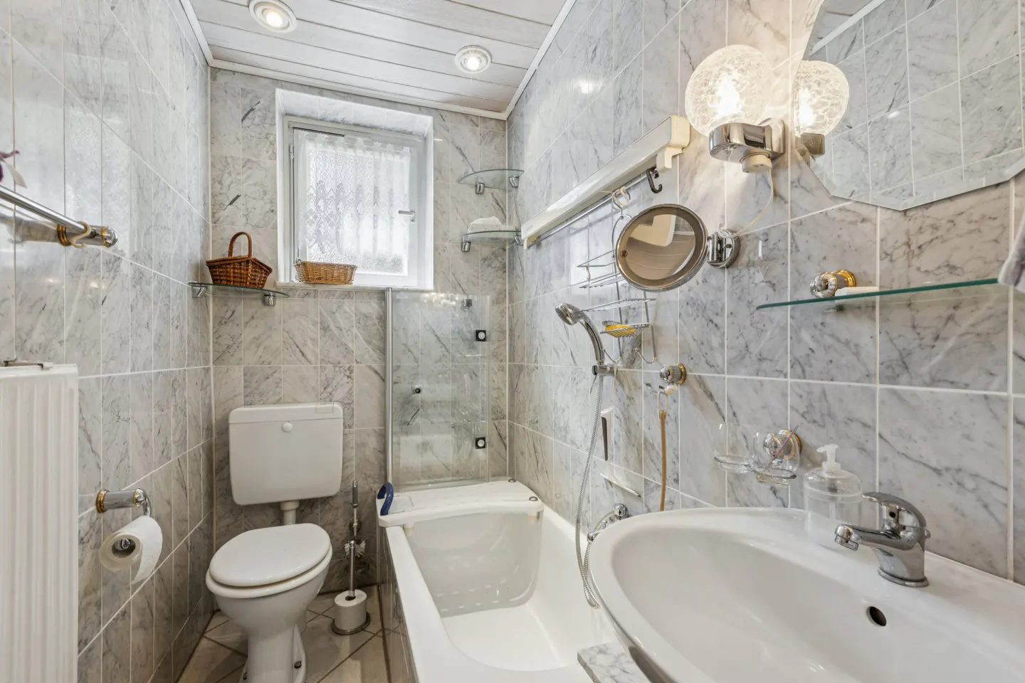 Bathroom with marble tile, white toilet, tub with glass door, and sink. A window with a lace curtain is above the toilet.