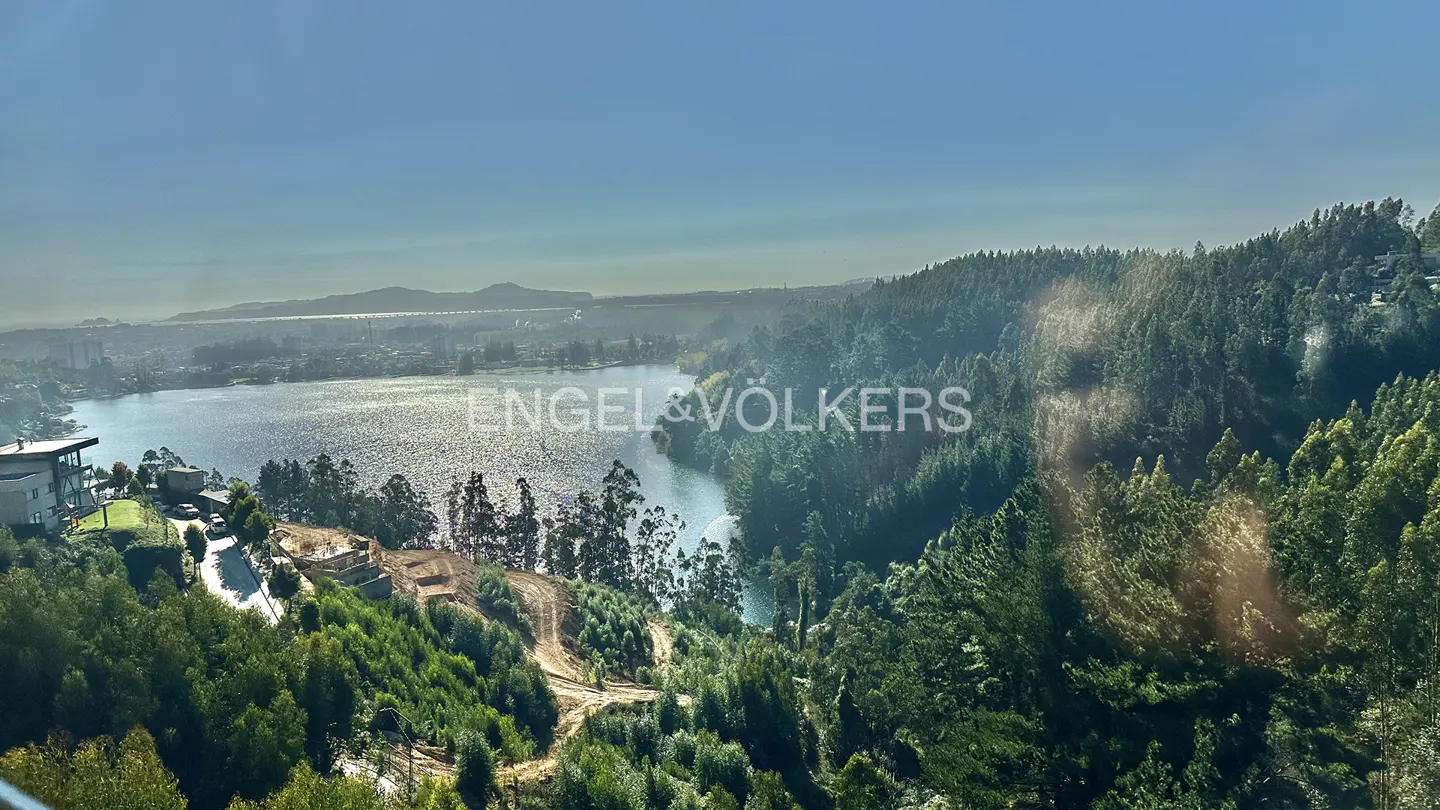 Scenic view of a lake surrounded by green trees and hills under a clear blue sky. Engel & Völkers logo is superimposed on the image.