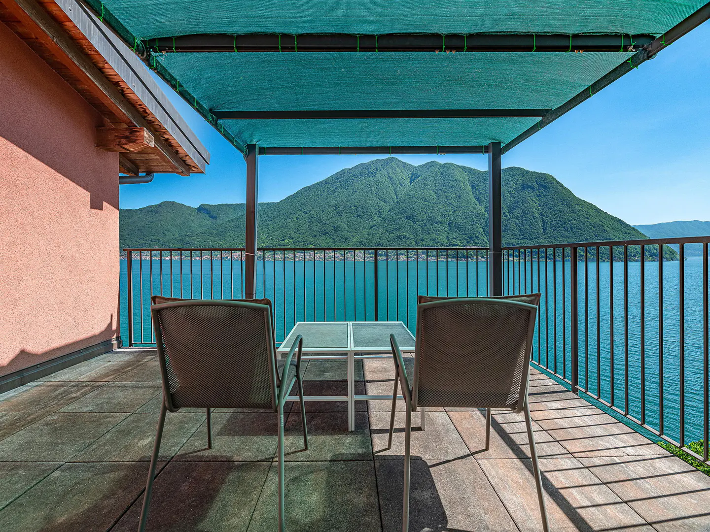 Balcony view with two chairs and a table overlooking a lake and mountain range on a sunny day.